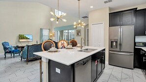 A modern kitchen with a central island, stainless steel appliances, and a dining area with a blue chair and a table. - Ultimate 5Br Villas With Pool/Spa (1555FD) (Davenport)