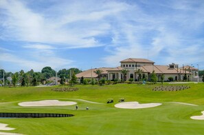 A golf course with bunkers, a clubhouse, and a large building in the background.