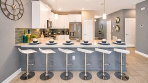 A modern kitchen with white cabinets, a central island with bar stools, and a large wall clock. - Luxury 9Beds Villa With Private Pool/Spa(1671MV) (Davenport)