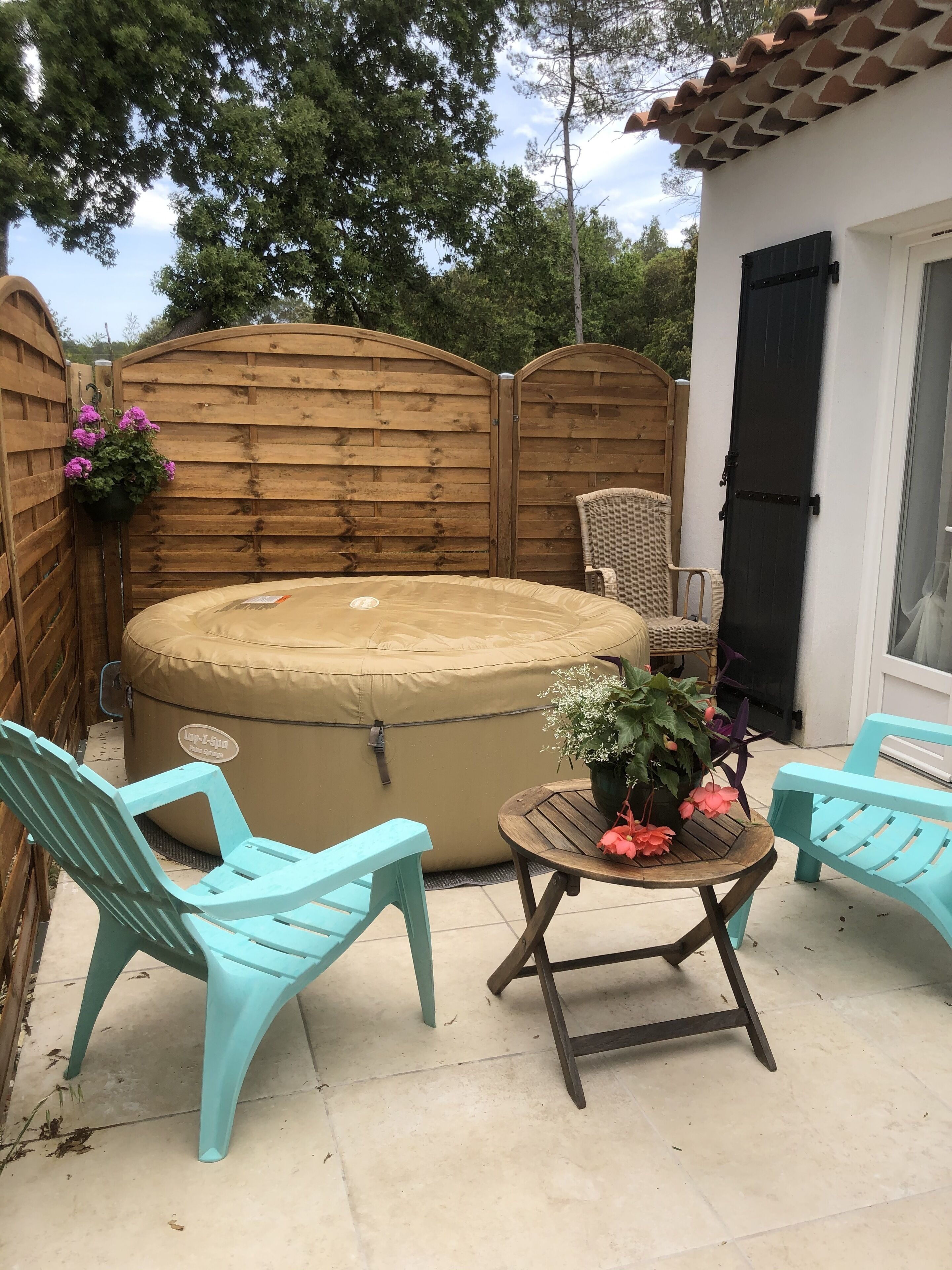 A patio area with a hot tub, two turquoise chairs, a round wooden table with a potted plant, and a wicker chair.