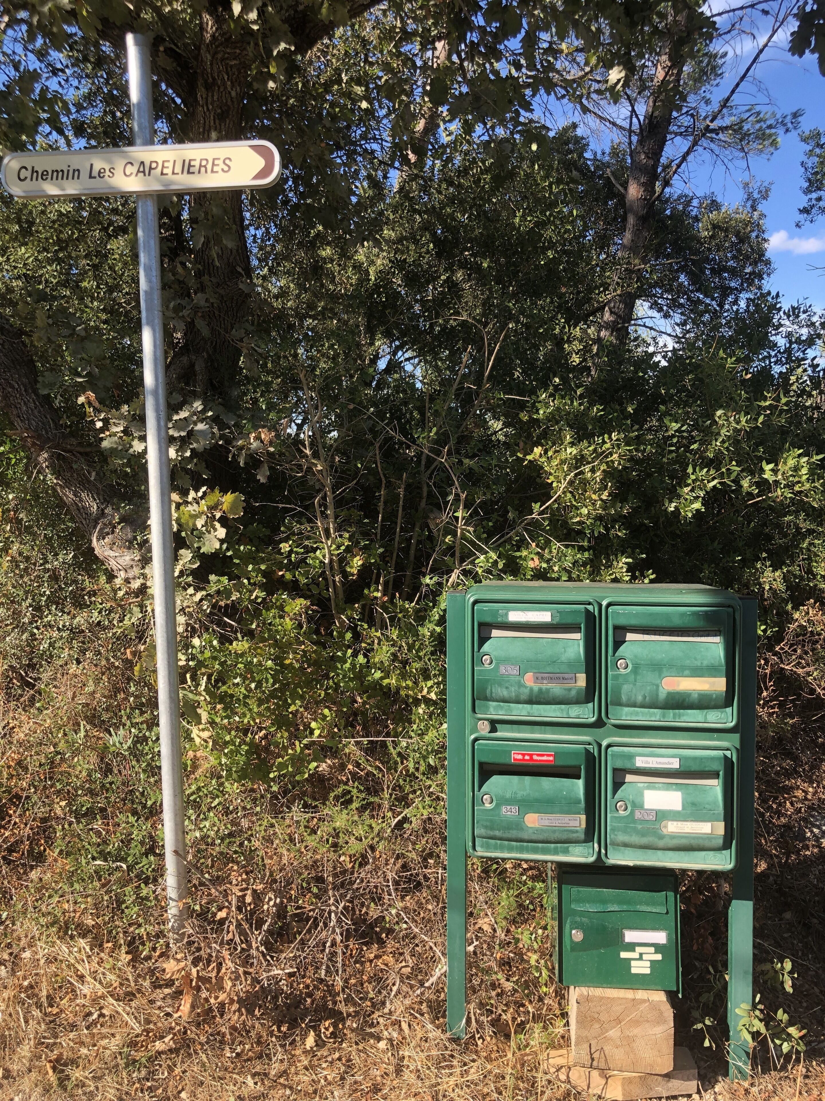 Ein Wegweiser für 'Chemin Les CAPELIERES' und ein grüner Briefkasten mit mehreren Fächern.