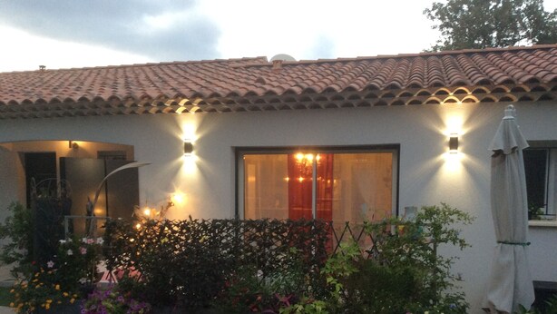 Una casa amb teulada de teules, dues finestres amb il·luminació exterior, una porta d'entrada i un jardí amb diverses plantes.