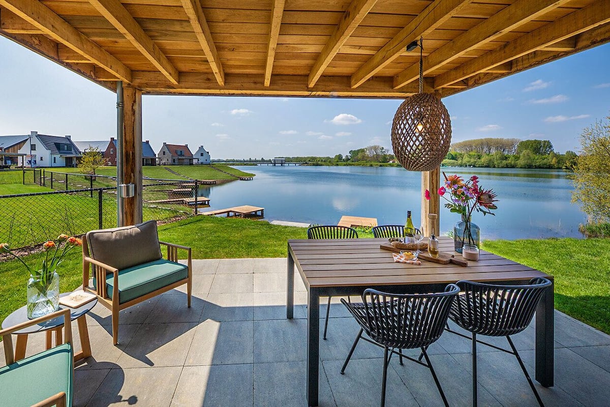 Une terrasse couverte, aménagée avec du mobilier d’extérieur, une table ornée d’un vase de fleurs, et une vue sur une rivière et des maisons en arrière-plan.