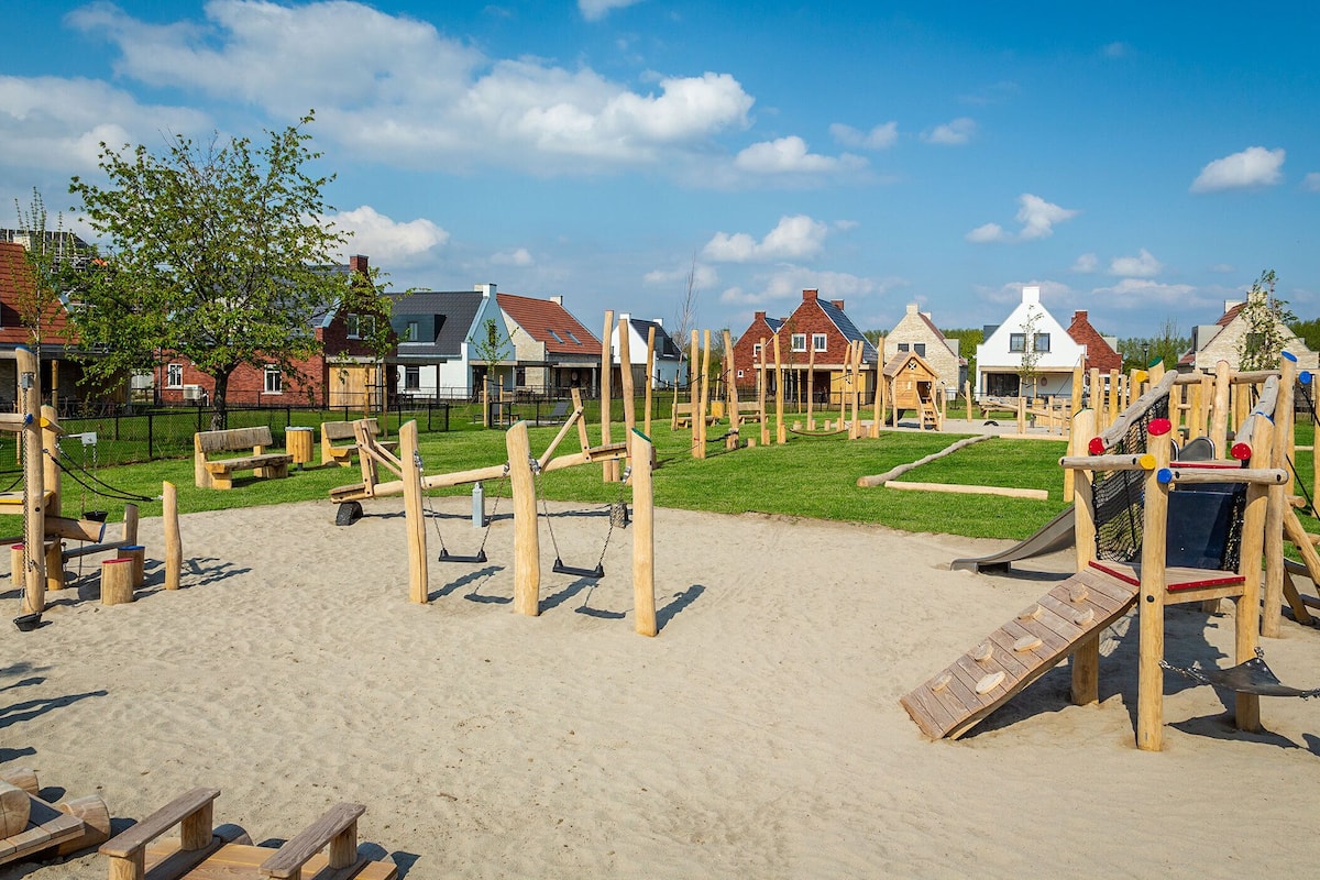 Un terrain de jeux doté de diverses structures en bois et d’une zone sablonneuse, entouré de maisons aux styles architecturaux variés.