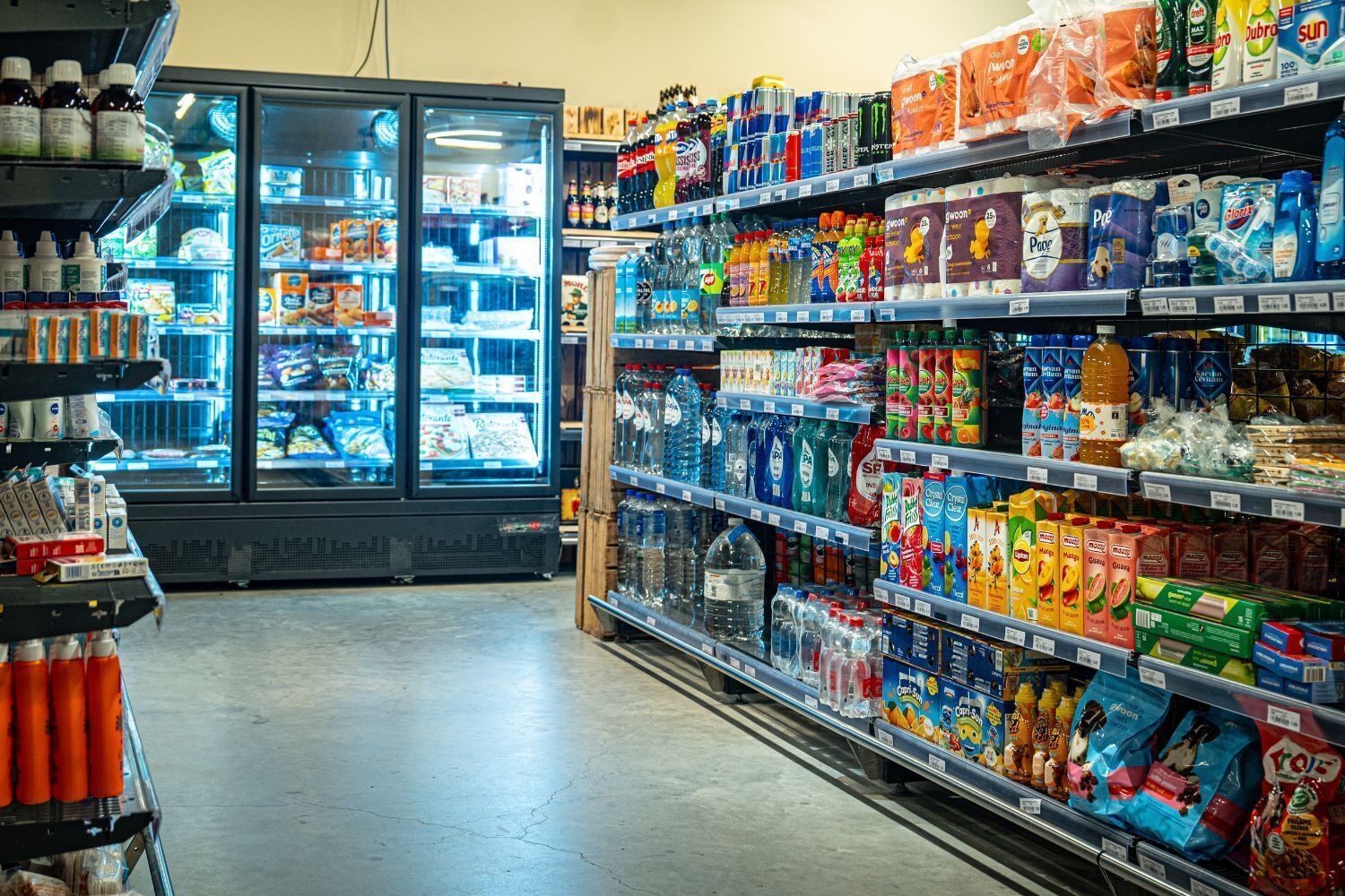 A grocery store aisle with refrigerated sections and shelves stocked with beverages, snacks, and household items.