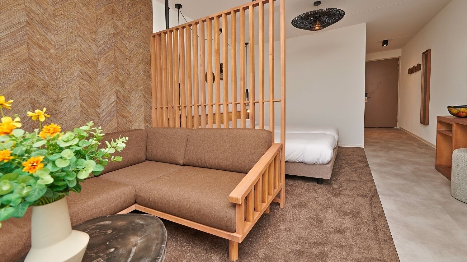 A modern living room with a sofa, a wooden partition, a small table with a vase of flowers, and a bed in the background.