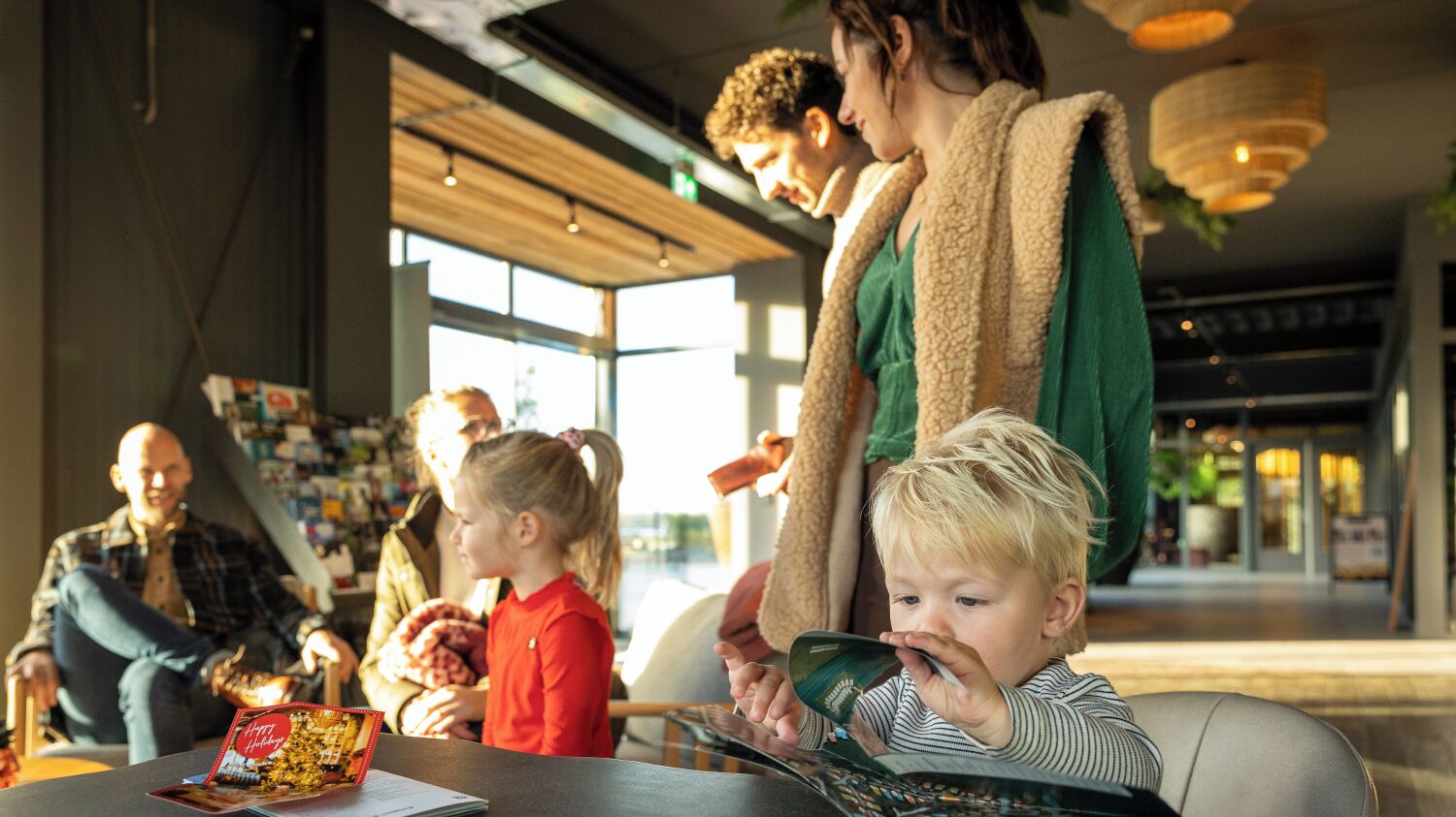 Una familia en un ambiente interior moderno con ventanas amplias, una mesa con una revista y una persona de pie con un niño.