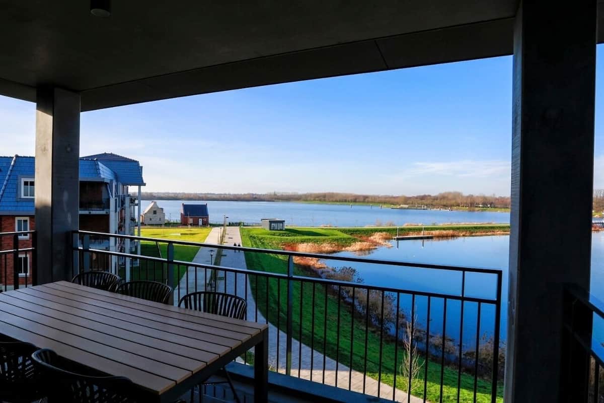 Un balcon avec une table et des chaises en bois, donnant sur un lac, avec un sentier et des bâtiments visibles au loin.