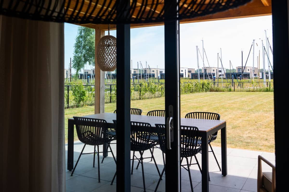 Une terrasse extérieure avec une table et des chaises, un luminaire suspendu et une vue sur un espace verdoyant, avec des bâtiments en arrière-plan.