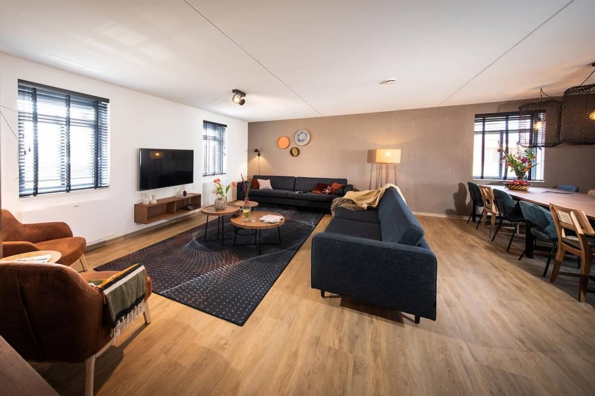 Un salon moderne avec un canapé, une table basse et un téléviseur. Il y a des fenêtres avec des stores, un coin repas avec des chaises et un coin cuisine avec une table et des chaises.