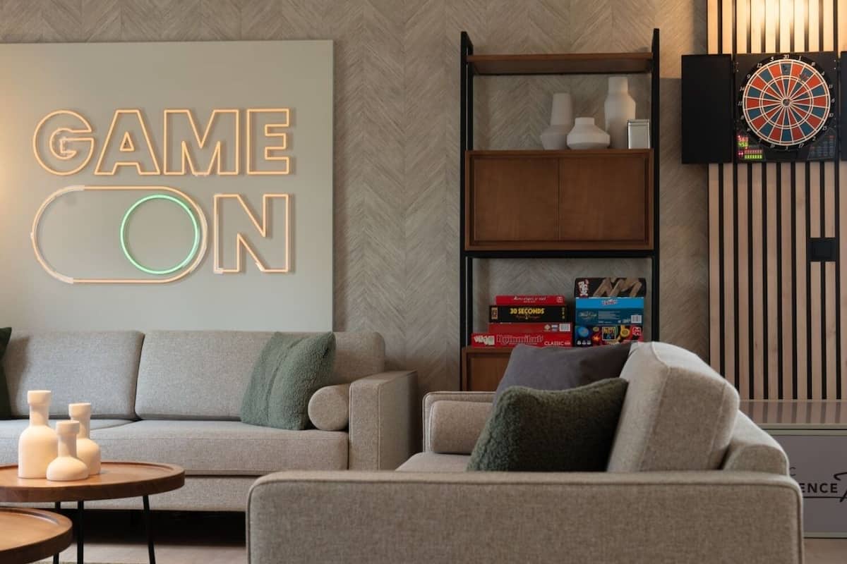Un salon moderne avec un canapé, une table basse et une bibliothèque. On y trouve un panneau néon sur lequel est écrit « GAME ON » et un jeu de fléchettes accroché au mur.