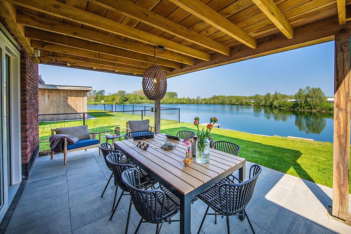 Une terrasse extérieure avec une table et des chaises en bois, un luminaire suspendu et une vue sur un lac.