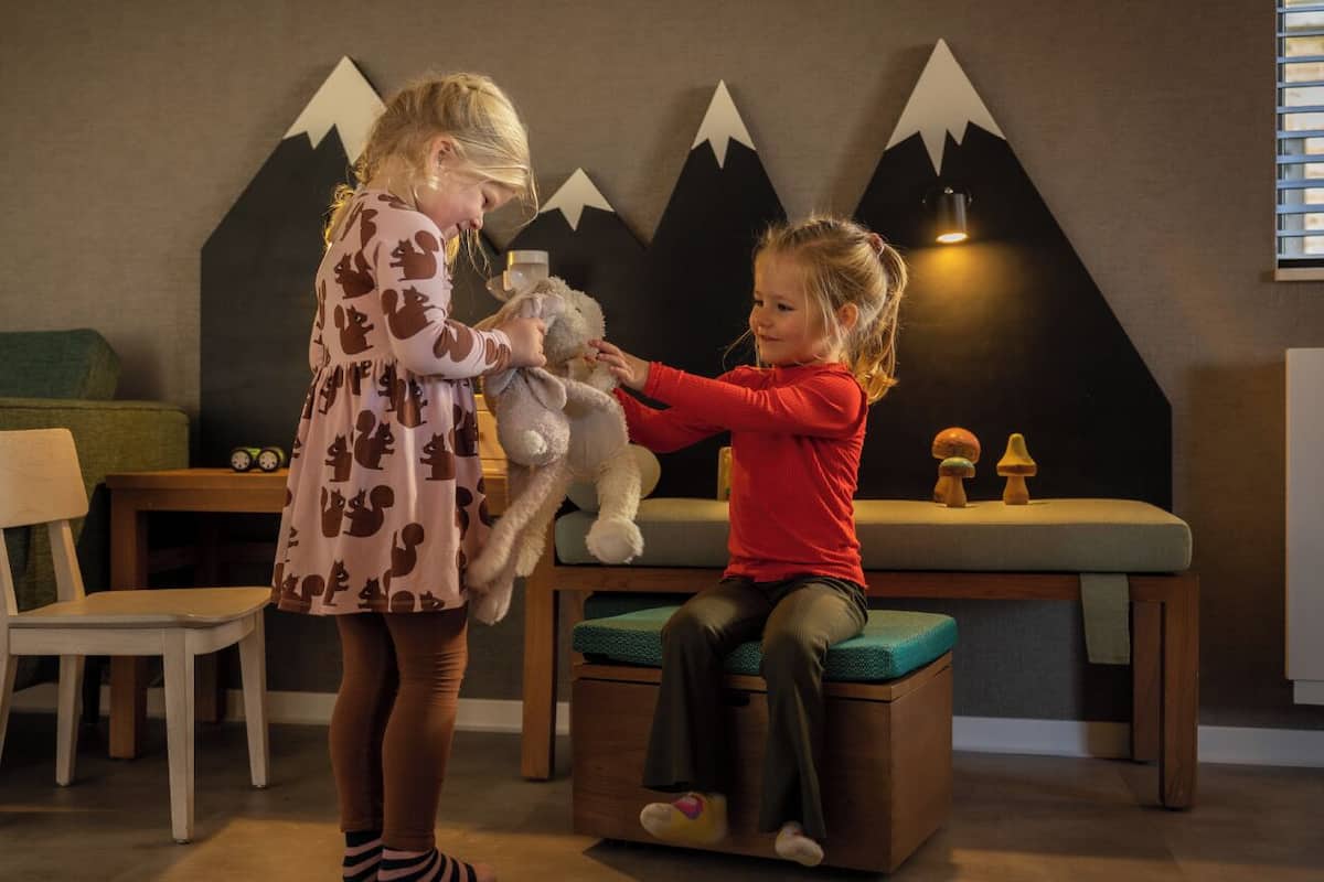 Deux enfants jouent avec un animal en peluche dans une pièce dont un mur est décoré sur le thème de la montagne, avec un canapé vert et une table en bois.
