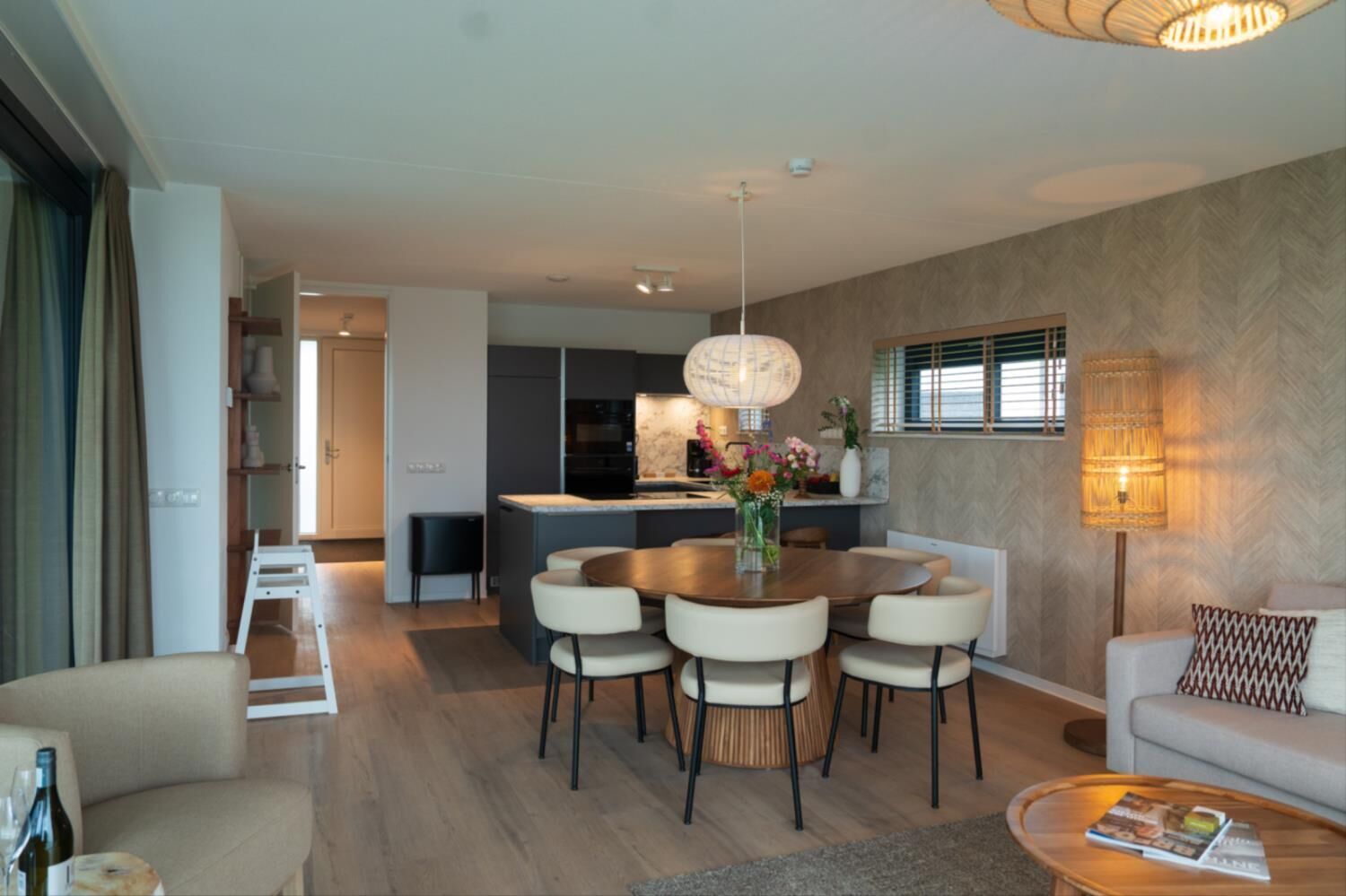 Ein modernes Wohnzimmer mit Essbereich, ausgestattet mit einem Holztisch, bequemen Sitzmöglichkeiten und einer Küche im Hintergrund.