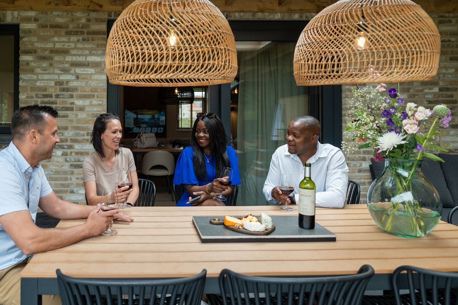 A group of people seated at a wooden table with a bottle of wine and a vase of flowers.