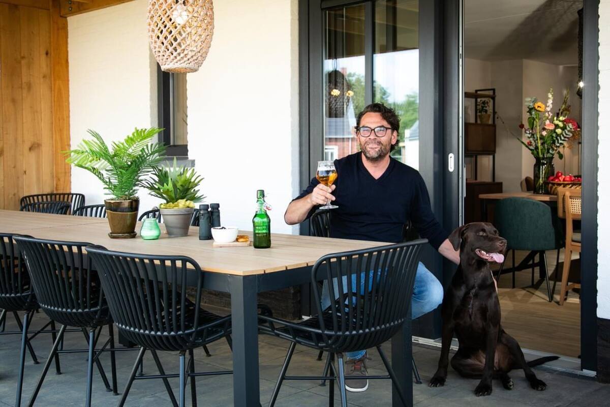 Un homme est assis à une table en bois, entouré de chaises noires, et tient un verre de bière. Un chien est assis à ses côtés.