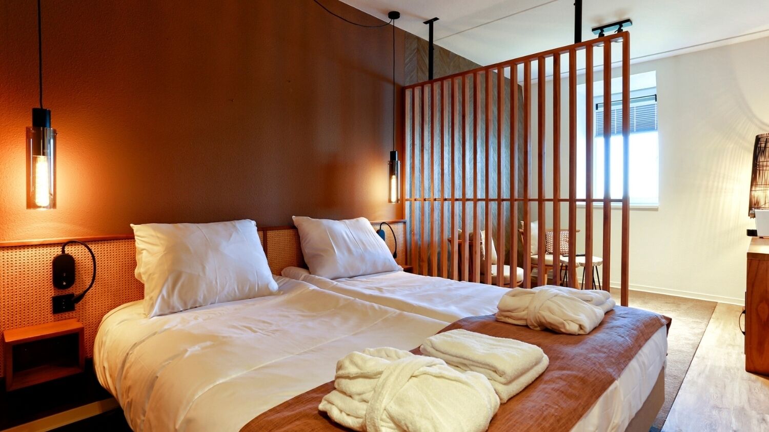 Hotelkamer met een groot bed, twee nachtkastjes, een houten hoofdbord, een scheidingswand en een raam met zonwering.