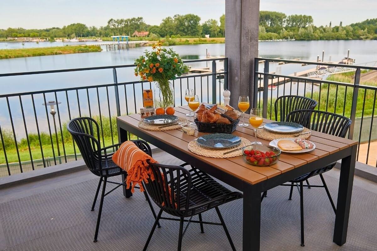 Une table à manger installée sur un balcon, d’où l’on peut admirer un lac, entourée de chaises et agrémentée d’un vase de fleurs.