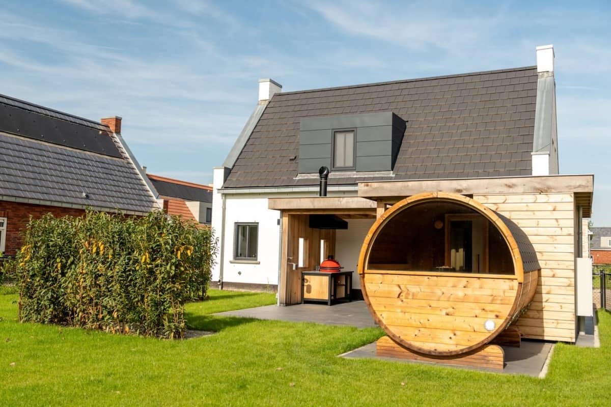 Une maison moderne avec un grand sauna en forme de tonneau en bois situé dans le jardin avant.