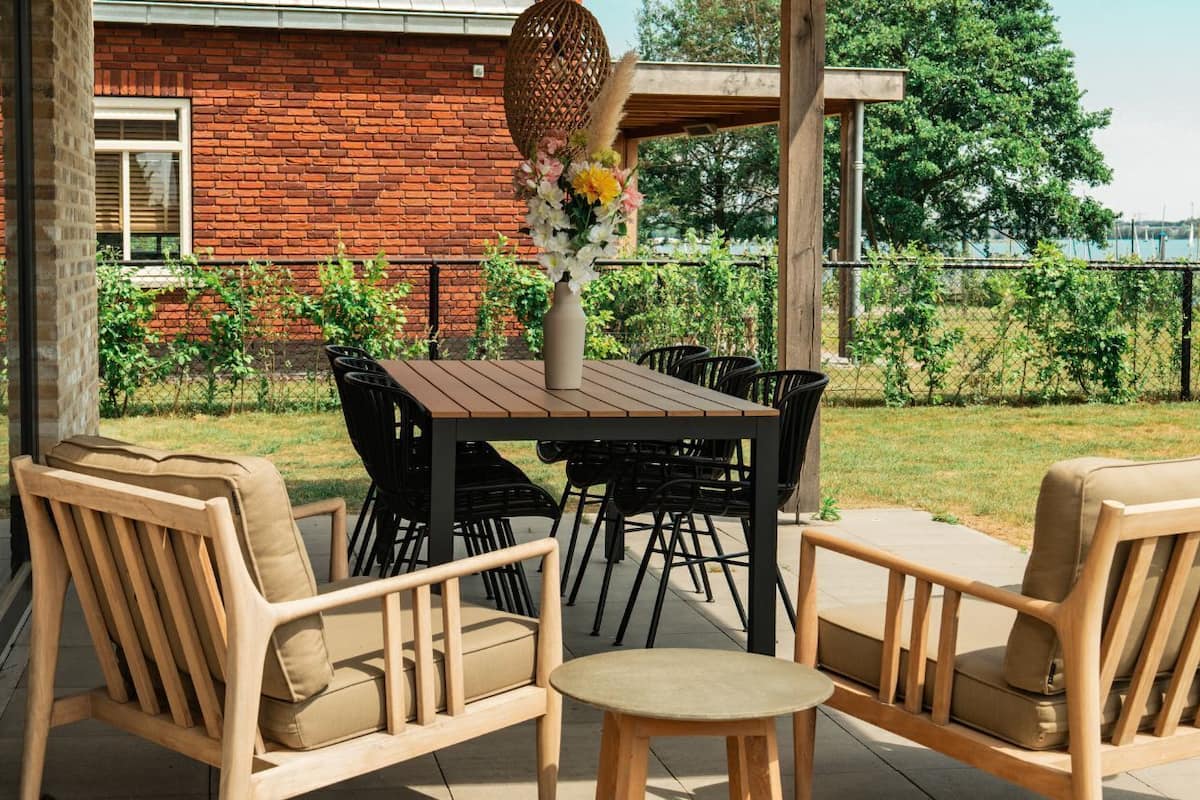 Une terrasse extérieure avec une table et des chaises en bois, un vase de fleurs et une vue sur un bâtiment en briques et sur la verdure.