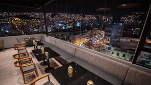 Un restaurante en la azotea con vistas a la ciudad, con mesas y sillas, y un área de estar con cojines.