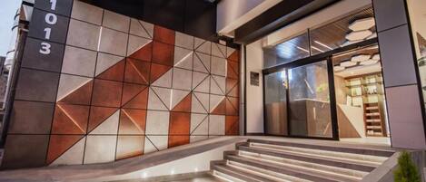 Una entrada moderna a un edificio con un muro de diseño geométrico, una escalera que lleva a la entrada y una puerta de cristal.