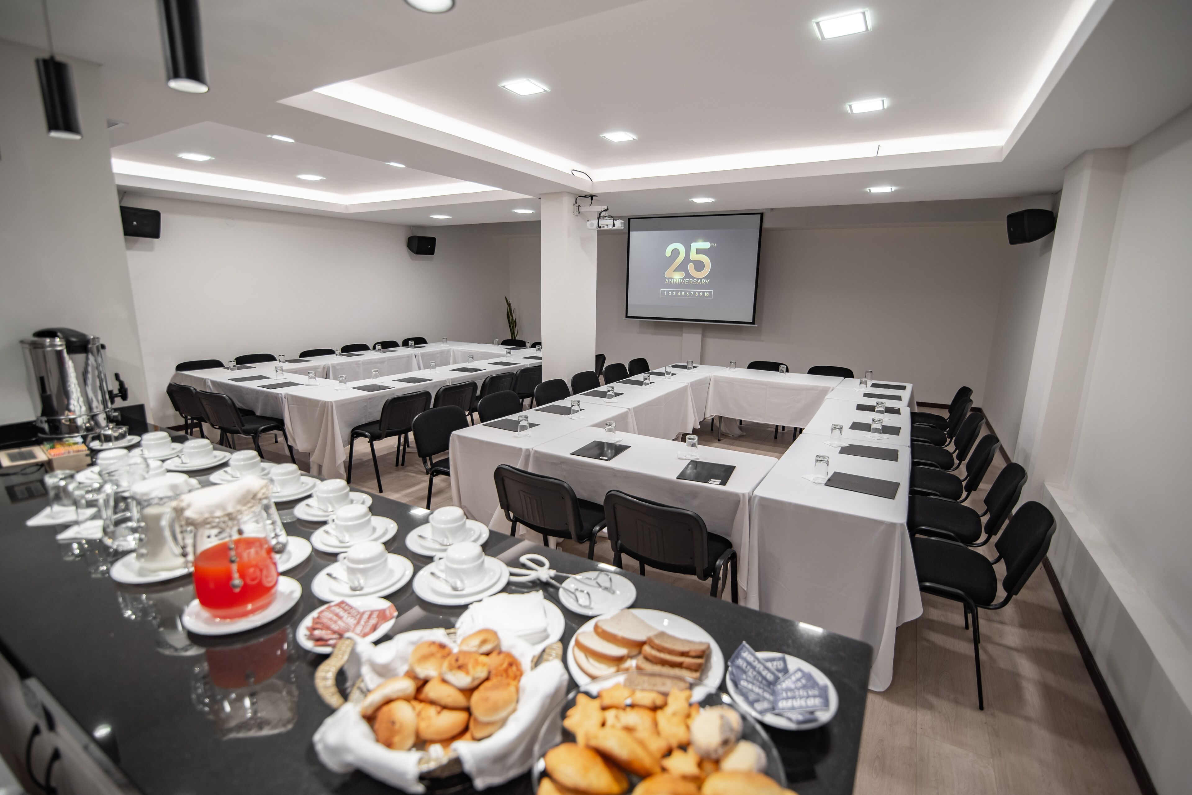 Una sala conferenze con un lungo tavolo apparecchiato per una riunione, provvista di sedie, uno schermo per proiettore che mostra '25 Anniversary' e un tavolo del buffet con diversi cibi.