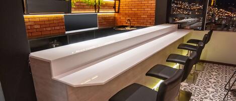 A modern bar area with a long counter, black bar stools, and a brick wall with shelves.