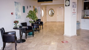A hotel lobby with a round table, chairs, a potted plant, a clock, and a television mounted on the wall.