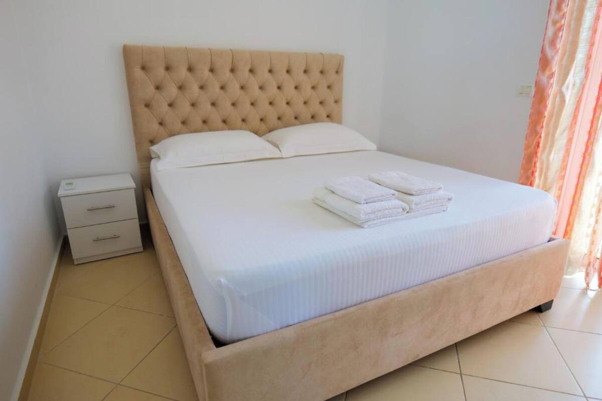 Una cama bien hecha con cabecera capitonada, una mesita de noche con cajones y una pared sencilla con una cortina a medio ver.