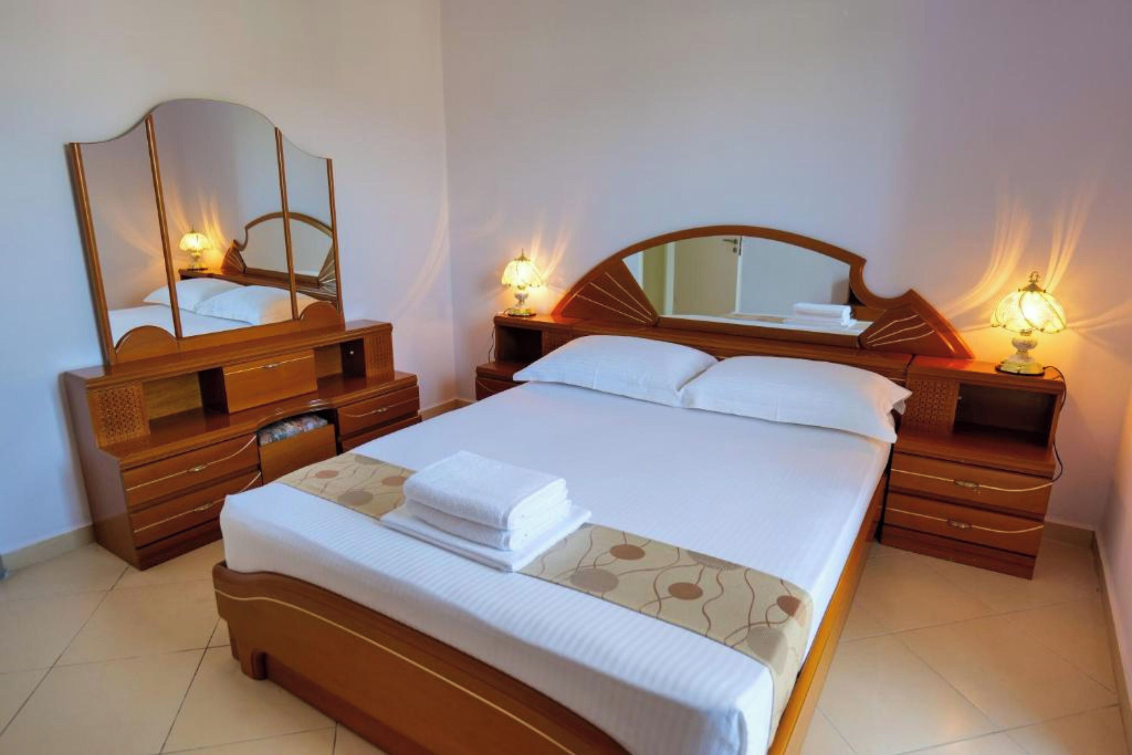 Una cama bien hecha con sábanas blancas y una toalla doblada, un aparador de madera con cajones, un espejo grande y dos lámparas de noche.