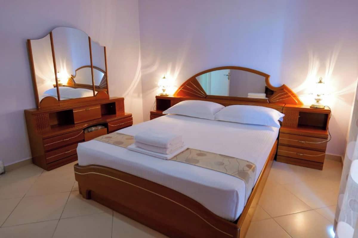 Una habitación de hotel con una cama grande, cabecera de madera, dos mesitas de noche con lámparas, un espejo y un aparador.