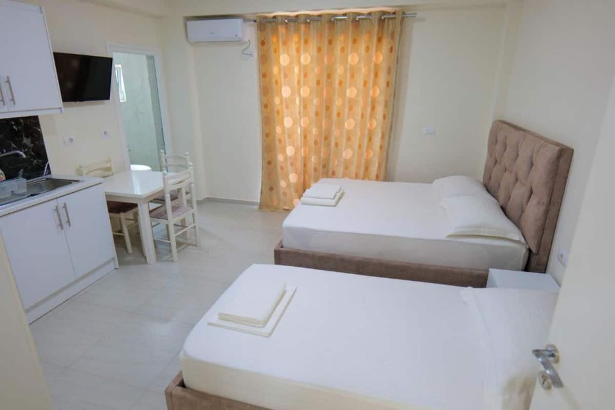 Habitación de hotel con dos camas, una kitchenette pequeña, una mesa de comedor y un televisor instalado en la pared.