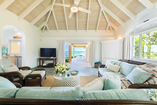 A spacious living room with a high ceiling, a television, wicker furniture, and a view of the ocean.