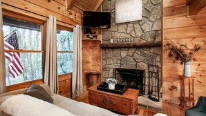 A cozy cabin interior with a stone fireplace, wooden walls, a television mounted above it, and a window with an American flag. - Sweet Dreams - Romantic and Hot Tub (Sevierville)