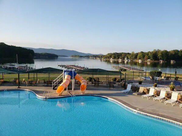 A swimming pool with a water slide, lounge chairs, and a view of a lake and hills.