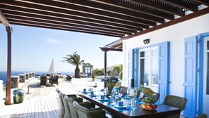 Un espace de restauration extérieur abrité, avec vue sur la mer, comprenant une terrasse en bois, des chaises en osier et une table dressée avec des serviettes et des assiettes bleues.