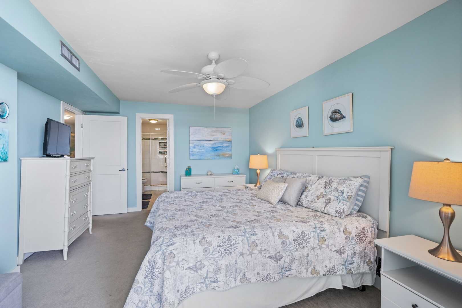 Une chambre à coucher comprenant un lit, des lampes de chevet, une commode, un ventilateur de plafond et une porte donnant sur une salle de bain.
