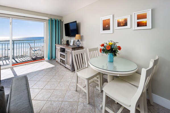 A balcony with a view of the sea, a dining area with white chairs and a table, and a TV mounted on the wall.