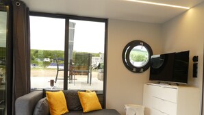 Ein modernes Wohnzimmer mit grauem Sofa, gelben Kissen, einem runden Spiegel, einem Flachbildfernseher und einem Couchtisch aus Holz.