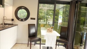 Eine moderne Küche mit Spüle, einem runden Spiegel und einem Fenster mit Blick auf eine grüne Landschaft. Zwei Stühle und ein kleiner Tisch mit einer Aufbewahrungsbox sind vorhanden.