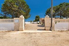 Une entrée fermée par un portail, sur laquelle on peut lire l’inscription « Dora », mène à un bâtiment à la façade en pierre, avec un ciel bleu clair en arrière-plan.