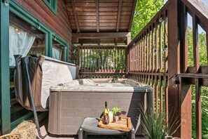 Une terrasse en bois avec un jacuzzi, une table avec une bouteille, des verres et une bougie, et une chaise avec une serviette.