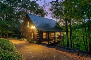 A wooden cabin with a sloped roof, surrounded by trees, and a paved driveway leading to the entrance.