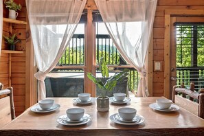 Une table à manger en bois, sur laquelle sont disposés six bols et soucoupes blancs, une plante en pot au centre, et des rideaux légers devant une fenêtre.