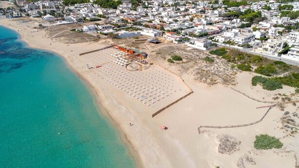 An aerial view of a coastal area with a beach, a row of white buildings, and a clear blue sea. - Villa Tina Maria by Perle di Puglia (San Pietro in Bevagna)