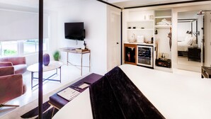 A modern hotel room with a large bed, a desk with a computer, a TV, and a glass partition separating the living area from the bedroom. - American Beech - King Suite 2 (Greenport)