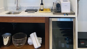 Une cuisine moderne équipée d’un évier, d’un plan de travail et d’un petit réfrigérateur. Des étagères au-dessus du plan de travail servent à ranger divers ustensiles de cuisine, et un tableau encadré y est également accroché.