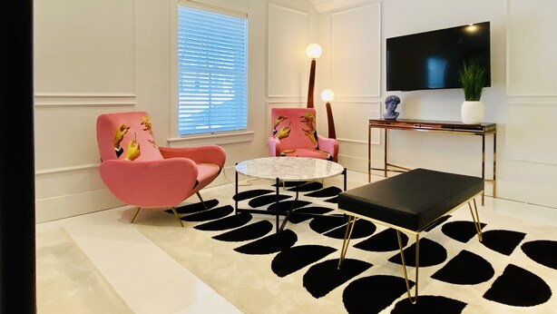 Un salon moderne avec deux fauteuils roses, une table basse noire, une table ronde blanche et un grand téléviseur à écran plat posé sur un meuble.