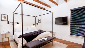 A four-poster bed with a bench, bedside tables, a flat-screen TV, and a window with blinds.